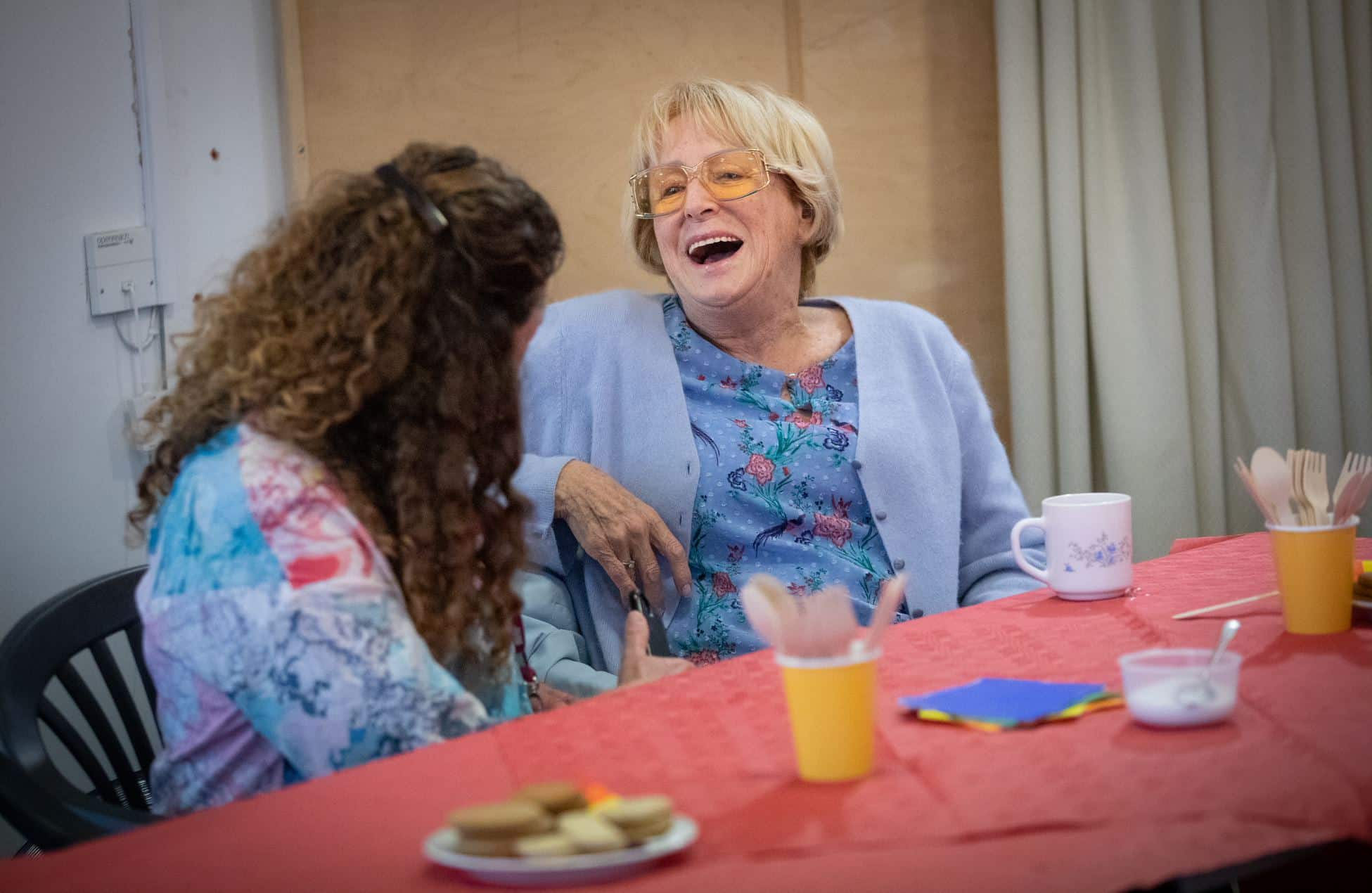 Two women sit at a table with biscuits and tea. One is facing away from the camera talking to an older woman who is laughing enjoying their chat.