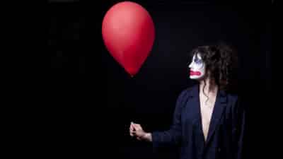 Laura Murphy looks at the red balloon she is holding. her face is covered in clown makeup and she wears a black jacket