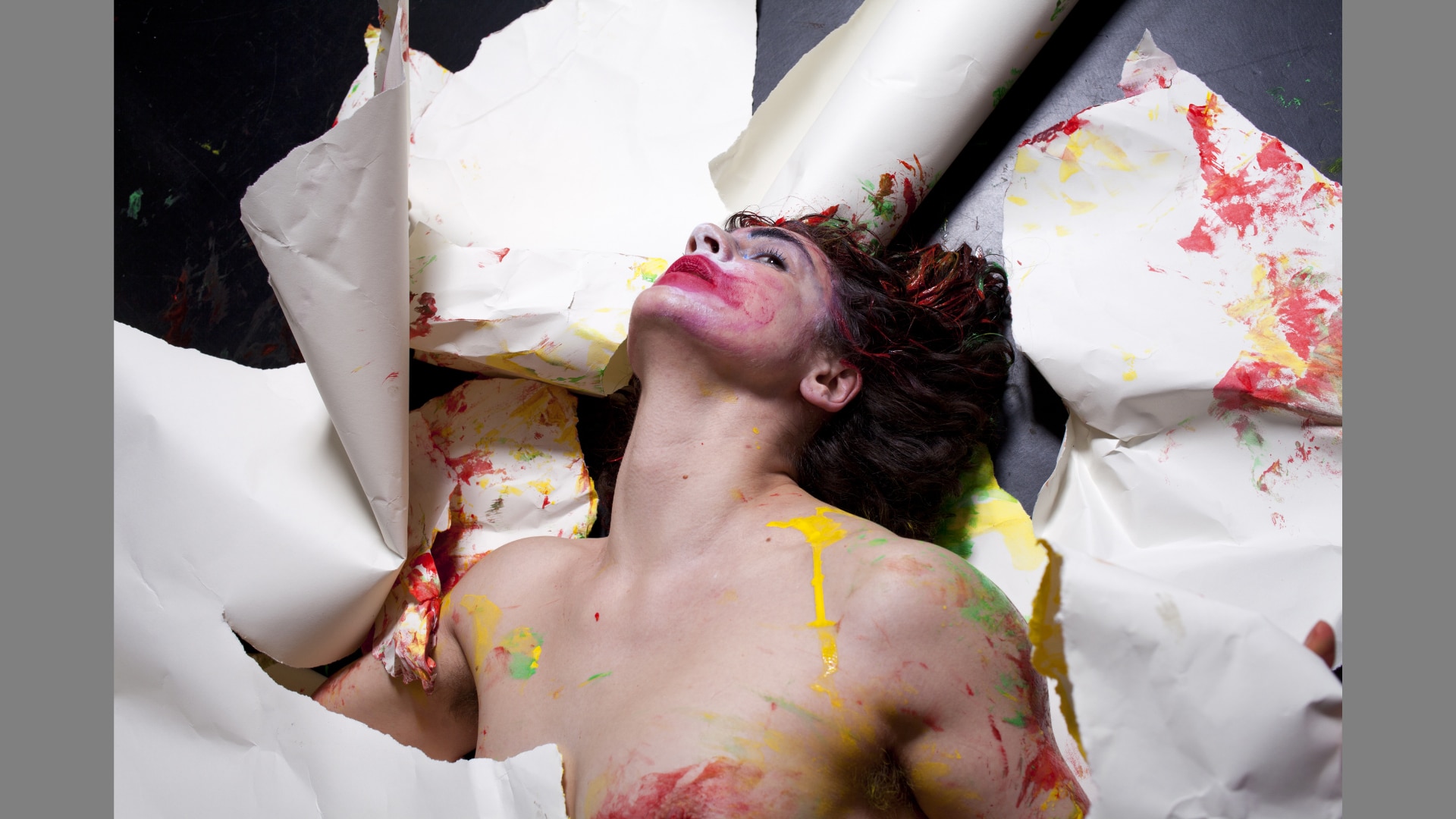 Laura Murphy bursts nude through masses of white material. She tilts her face back. Her face is covered in thick makeup