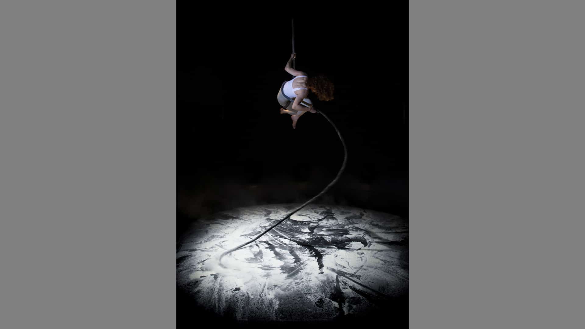 Photo of Rebecca Youssefi performing arial work high above the stage, where a pool of speckled white light under her