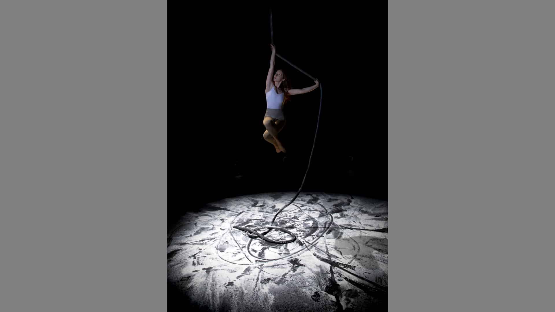 Photo of Rebecca Youssefi performing arial work high above the stage, where a pool of speckled white light under her