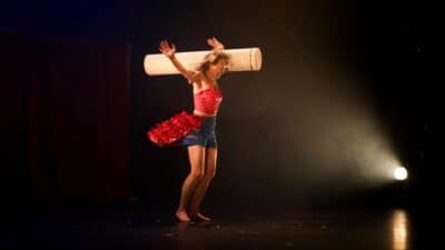 A woman stands on a stage, a white light shines on her from the right. She is spinning around with a cylindrical tube on her shoulder, she is laughing.