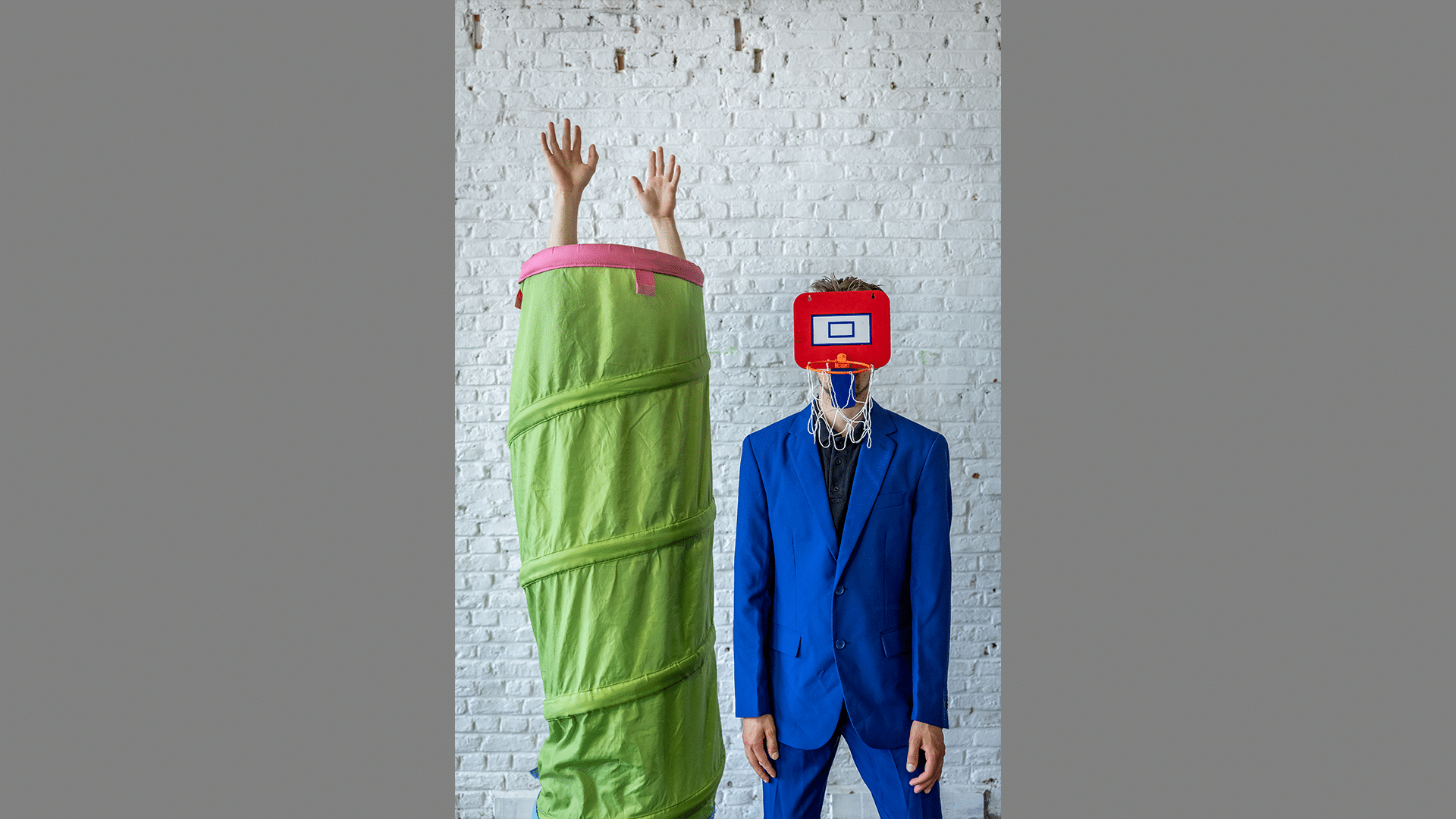 A performer in a blue suit has a mini basketball hoop covering his face, a second performer stands next to him covered up by a green collapsible tunnel.