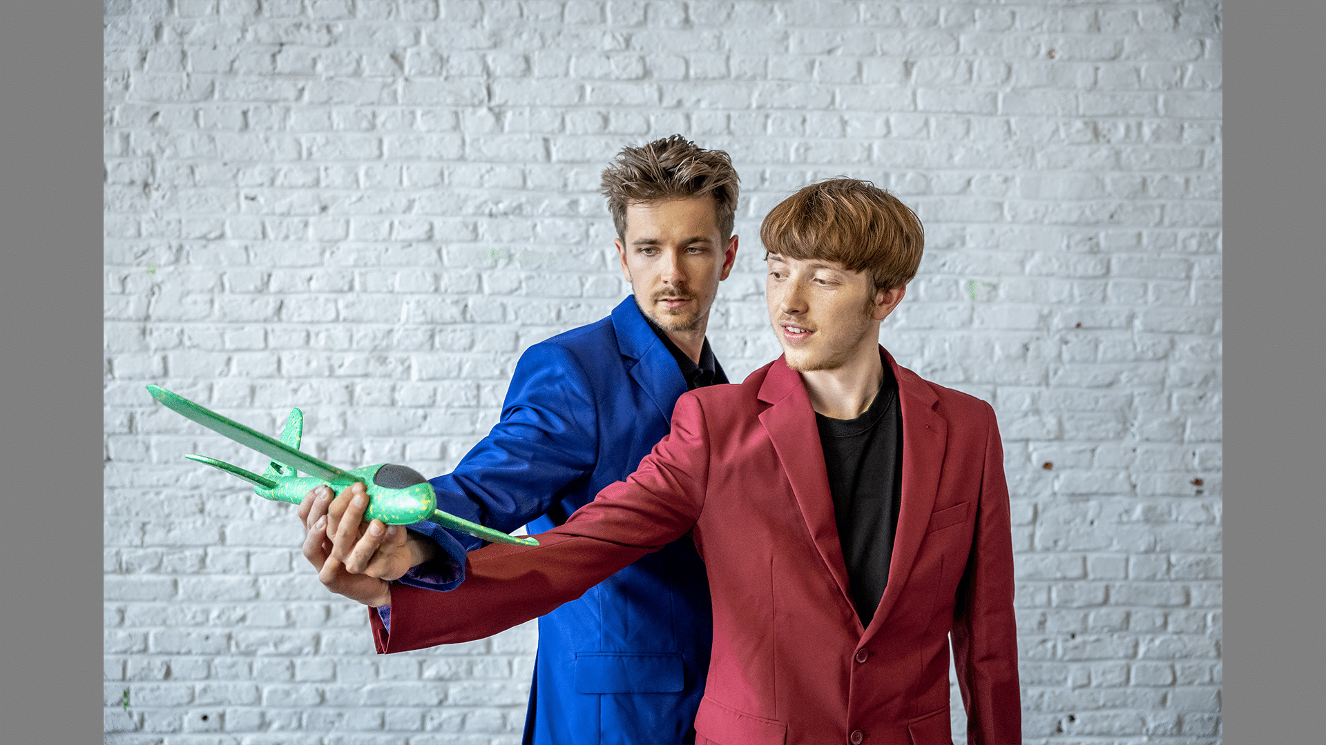 Two performers wear red and blue suits and hold a miniature plane.