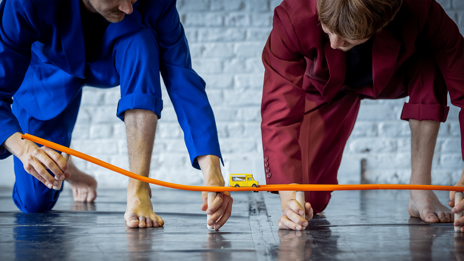 Two performers wearing red and blue suits balance a toy car track on their hands, a yellow toy bus rolls along it.