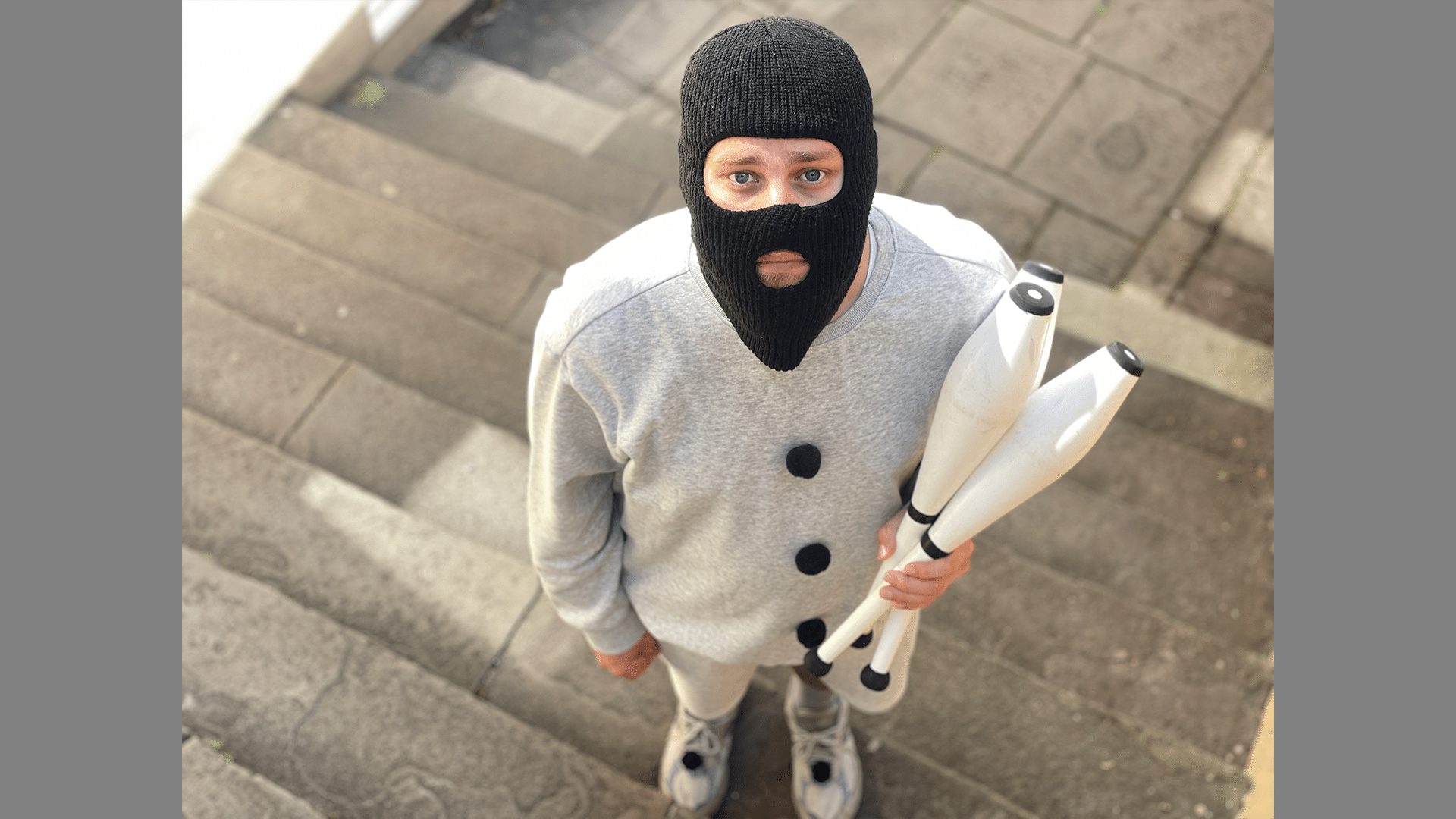 A performer in a balaclava looks up at the camera, they are holding juggling clubs.