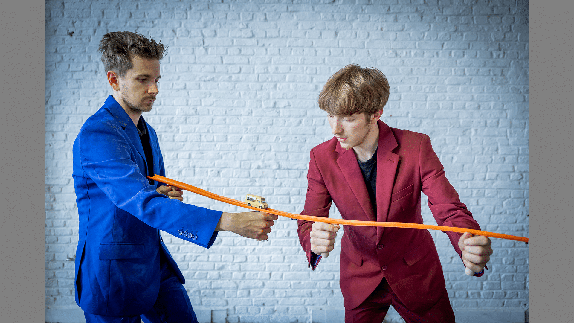 Two performers wearing red and blue suits balance a toy car track on their hands, a yellow toy bus rolls along it.
