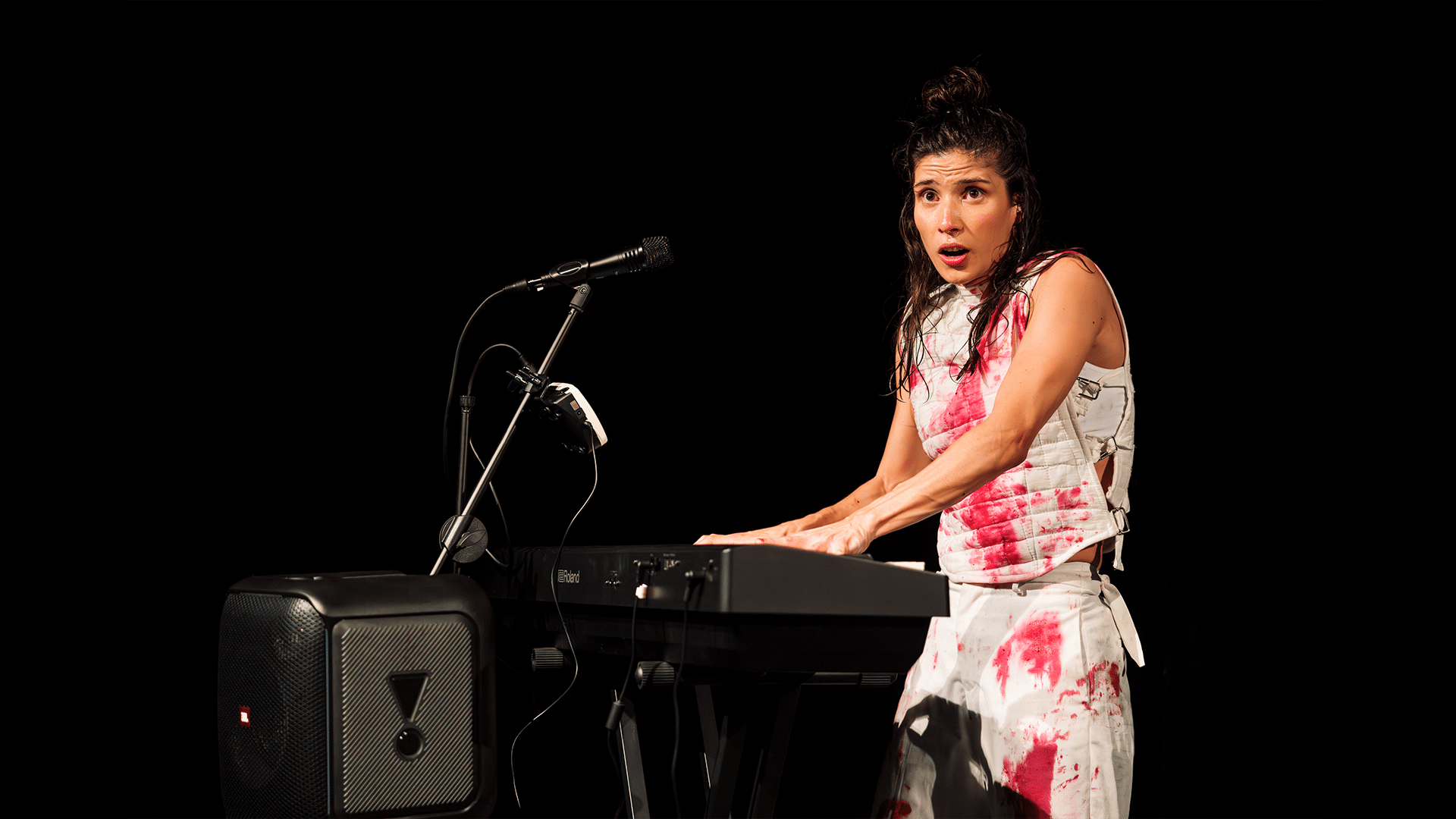 A woman wearing a white outfit covered in red stains plays the keyboard, she looks shocked.