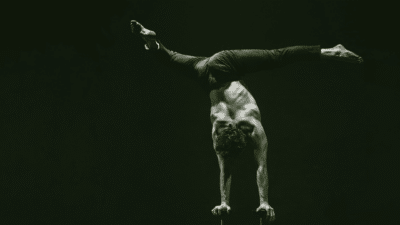 A handstand artist balances on their hands, their legs are in a split position. They are against a black background.