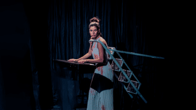 A performer stands against a navy backdrop, she has a stepladder around her shoulders and it falls behind her like a cape.
