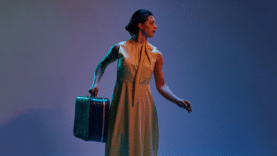 A woman stands against a purple background, she is wearing a cream coloured dress and holding a suitcase, she is looking to the left.