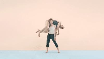 A women stands on a blue floor, set against a pale pink background. She holds a man horizontally around her shoulders.