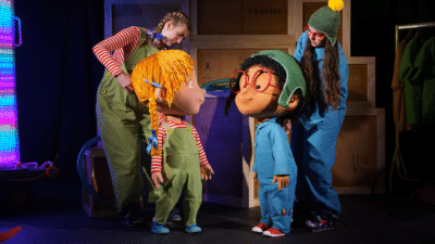 Two actors are on stage with two large puppets, one is a young girl with blonde hair in pigtails who wears a green dungaree and a red striped t-shirt. The other is a young person weaing a blue jumper and blue trousers, with red glasses and a green hat.