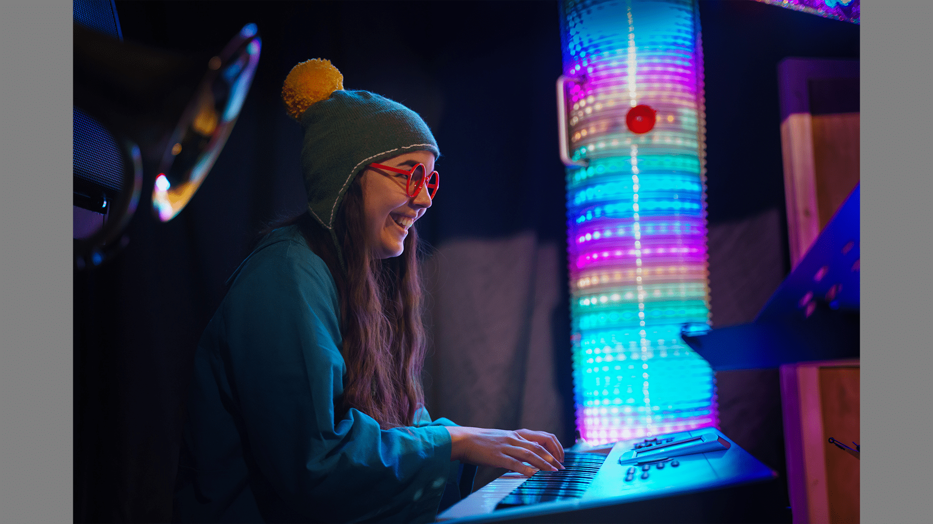 A woman plays keyboard, she has red glasses on, and a green bobble hat with a yellow bobble.