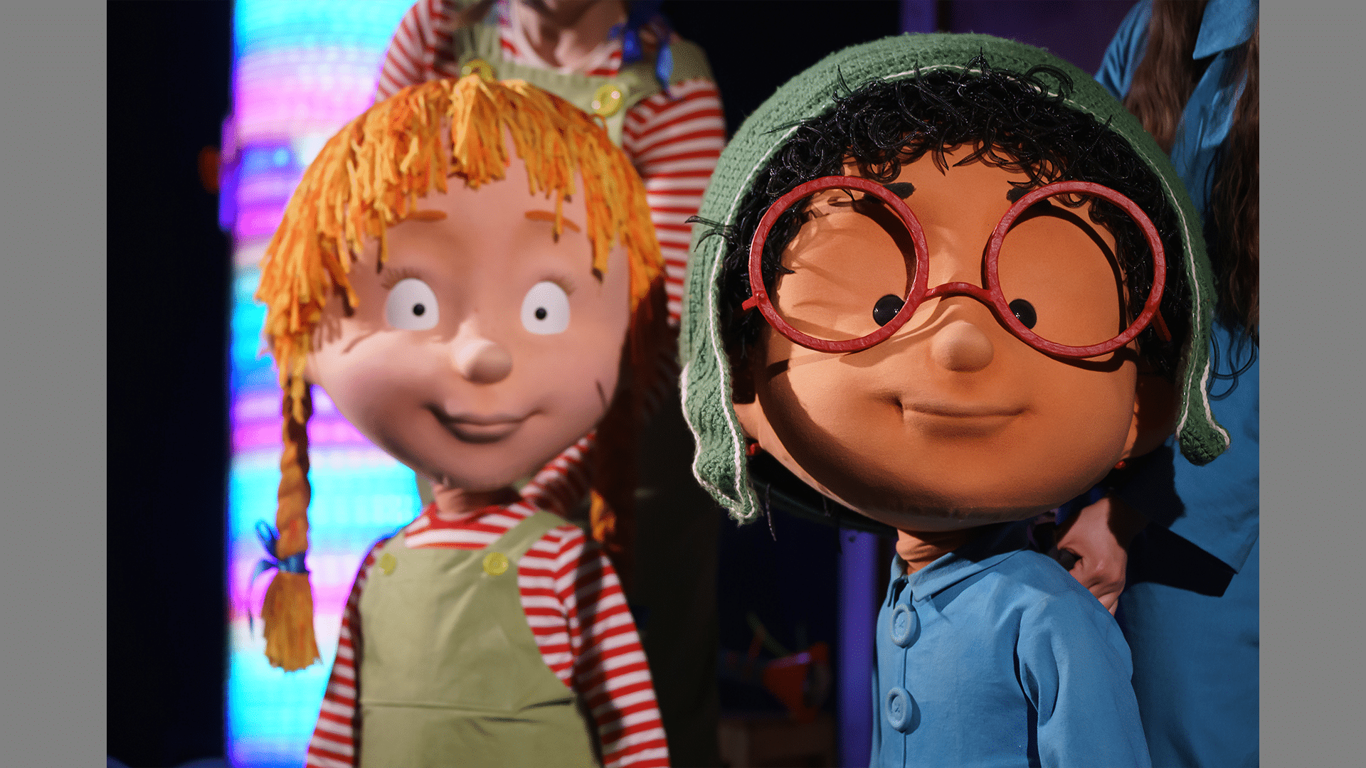 Tilly and Norbert stand looking at the camera. Tilly has ginger hair in two plaits, and Norbert has round red classes and a green hat.