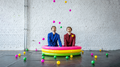 Two adults sit in a pink, yellow, and green inflatable swimming pool, they are throwing pink, yellow, and green balls out of the pool.