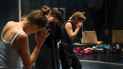 Three artists stand together in a rehearsal room, they have their hands to their faces as though shielding their eyes and are looking down.