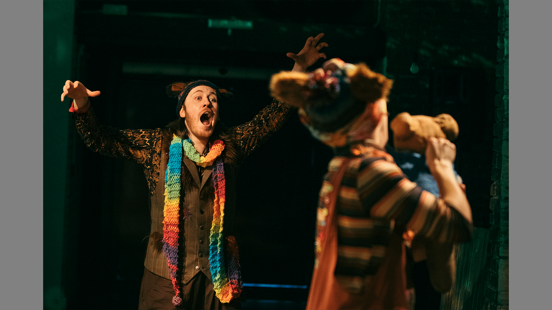 A performer stands on stage holding a bear puppet, their back is turned away from the camera. They are looking at another performer who wears a rainbow coloured scarf, it looks like he is jumping out on them.