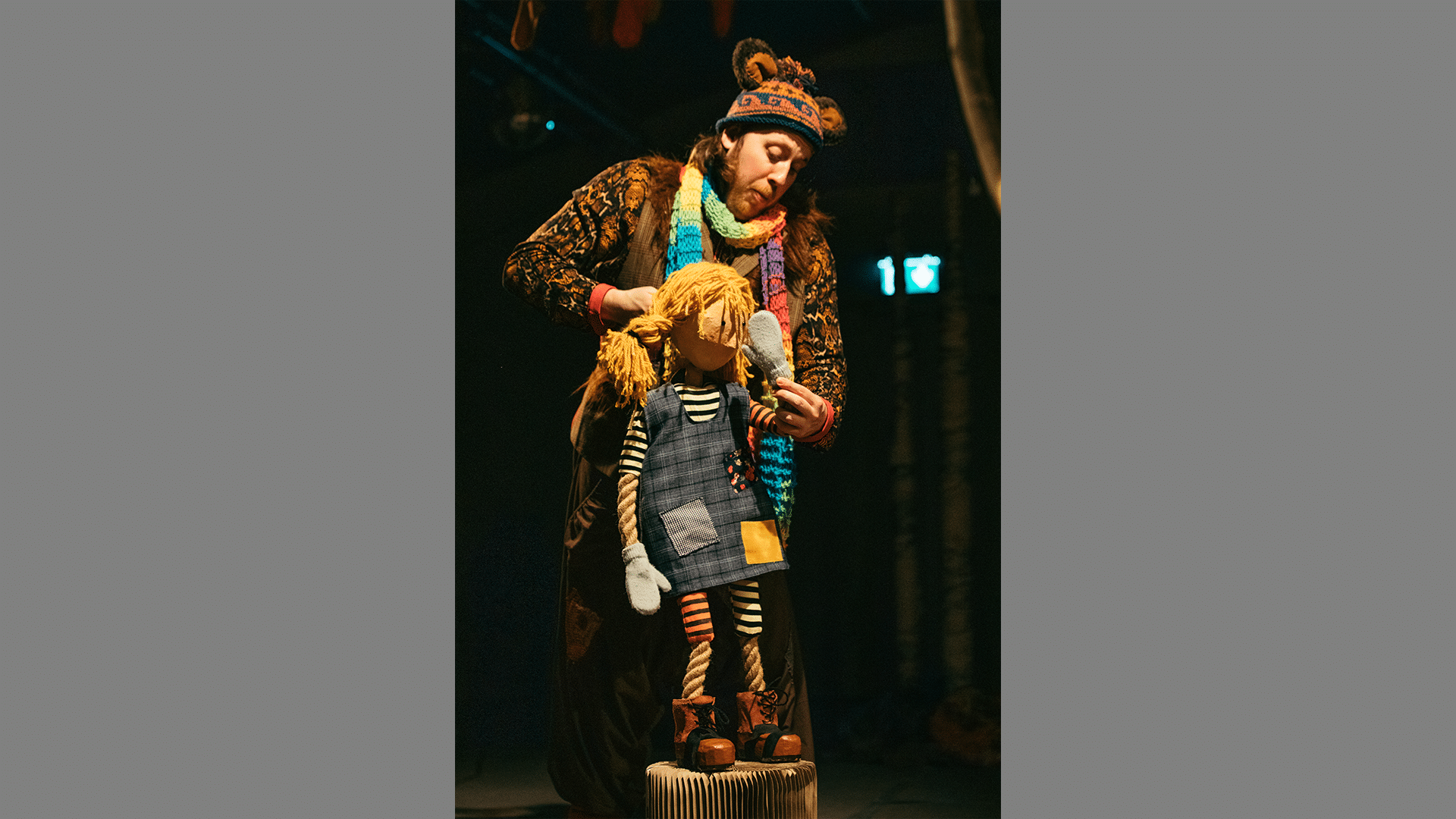 A performer stands on stage with a puppet of a young girl who has golden hair.