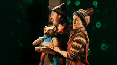 Two performers wearing knitted hats with bear ears on them stand against a green background, they are holding a little bear puppet.