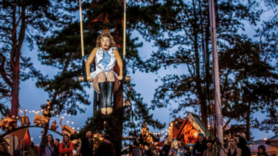A circus artist sits on a trapeze in the middle of a forest.