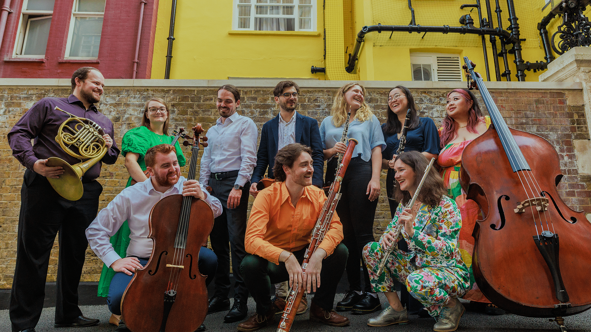 A collective of musicians smile at each other, they are holding various musical instruments including a bassoon, double bass and flute.