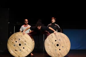 A rehearsal image. A performer balances themselves on two cable reels , two other performers standing back.
