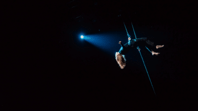 A person lies back whilst in midair in a harness. A blue spotlight shines on them.