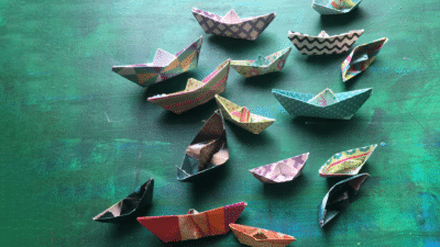 Origami boats made from lots of different patterned paper lie on a green tabletop.